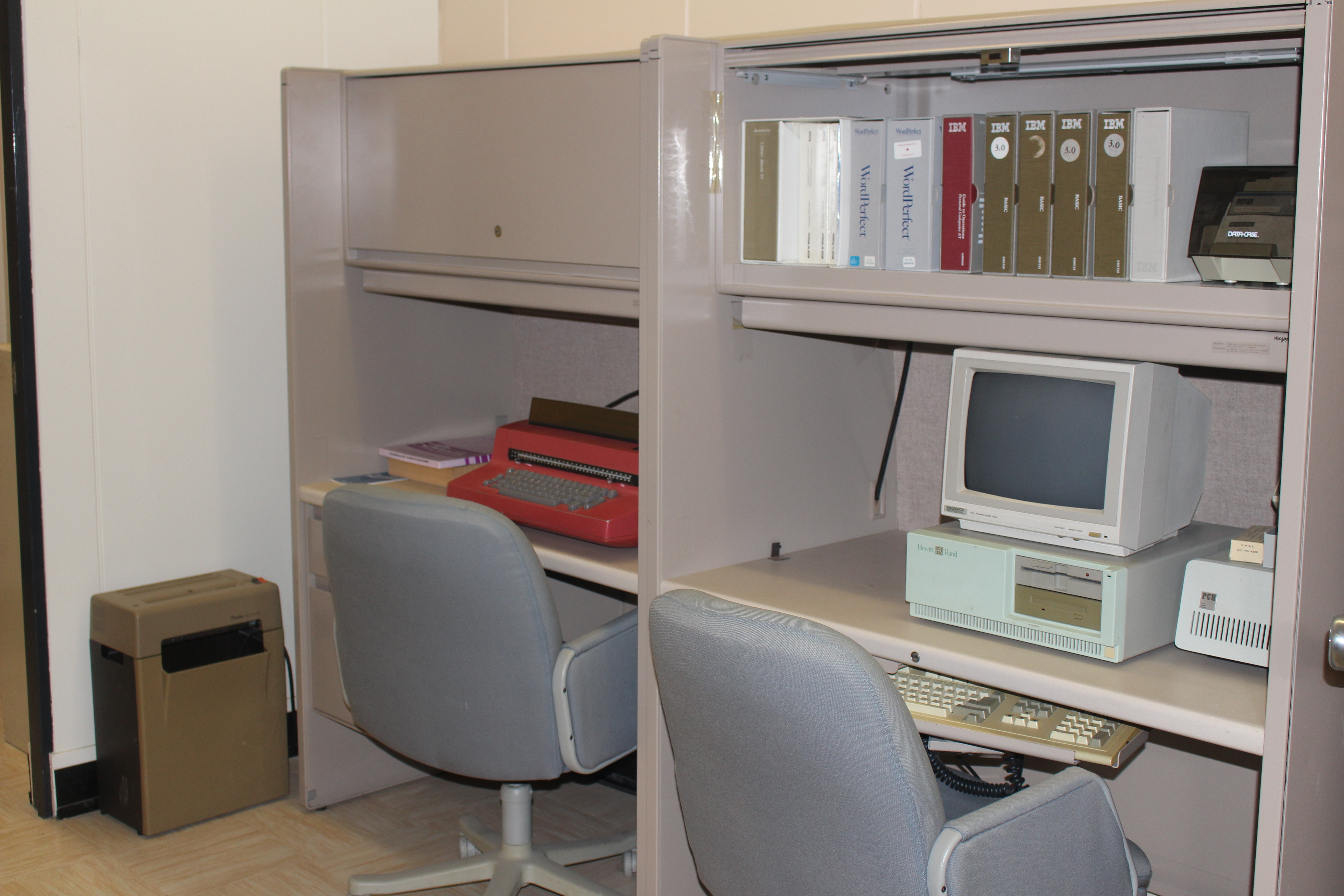 Office cubical with a typewriter and computer on the desk and a paper shredder on the ground. 