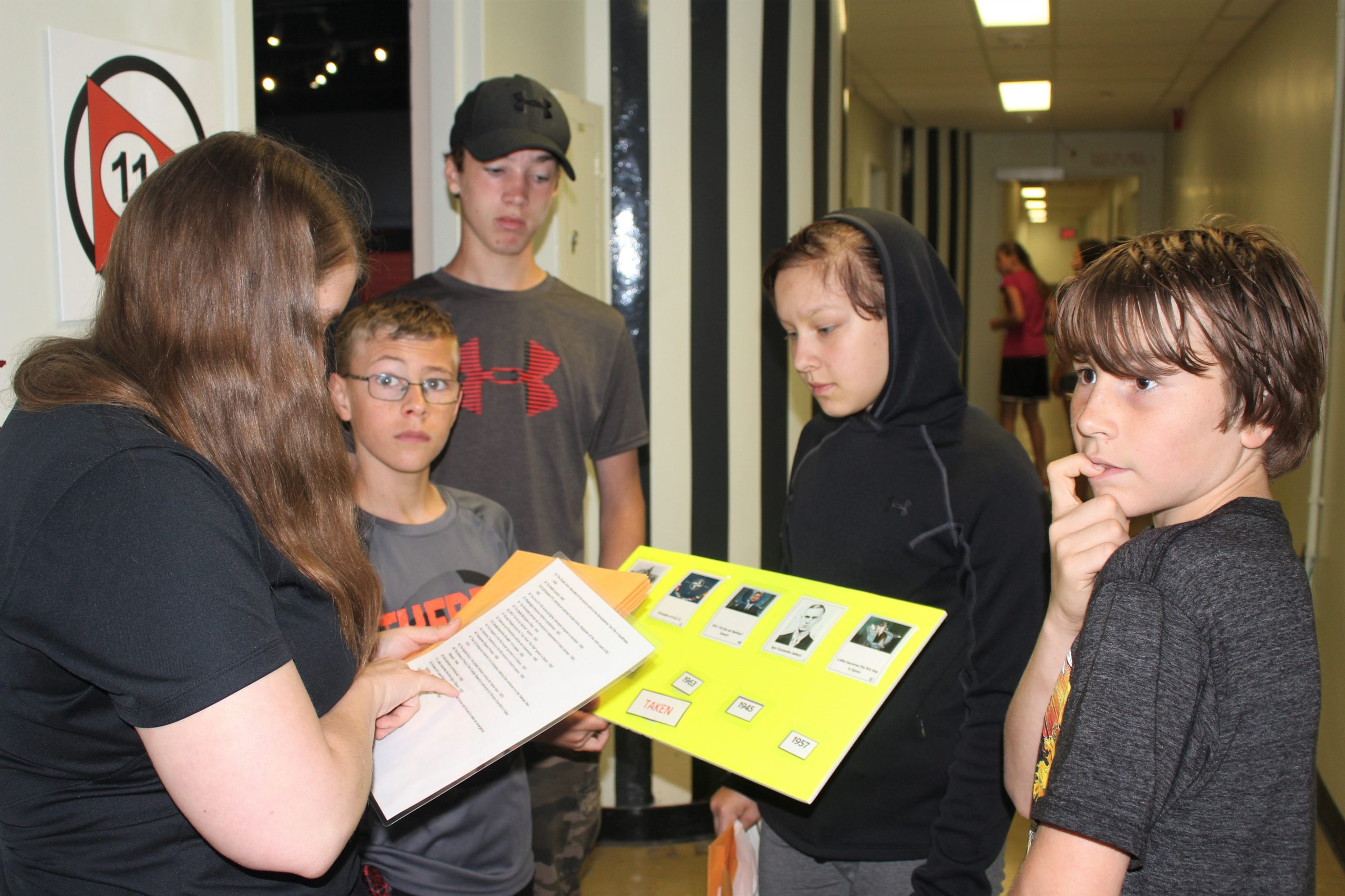 Four students look at a board of clues held by the facilitator in front of them in the hallway of the Diefenbunker.
