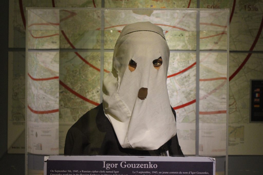Image of Igor Gouzenko wearing a face covering with a map behind him and his name plate on the desk in front of him.
