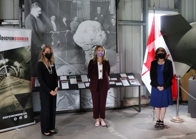 Diefenbunker Executive Director Christine McGuire, MP for Kanata-Carleton Jenna Sudds, and Diefenbunker Board Vice President Joanne Charettestand in front of an exhibit at the entrance to the Diefenbunker.