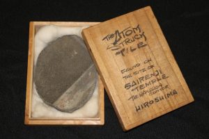 The Atom Struck Tile nestled in white fabric placed in the centre of a wooden box that reads "The Atom Struck Tile found on the site on Sairenji Temple Hiroshima."