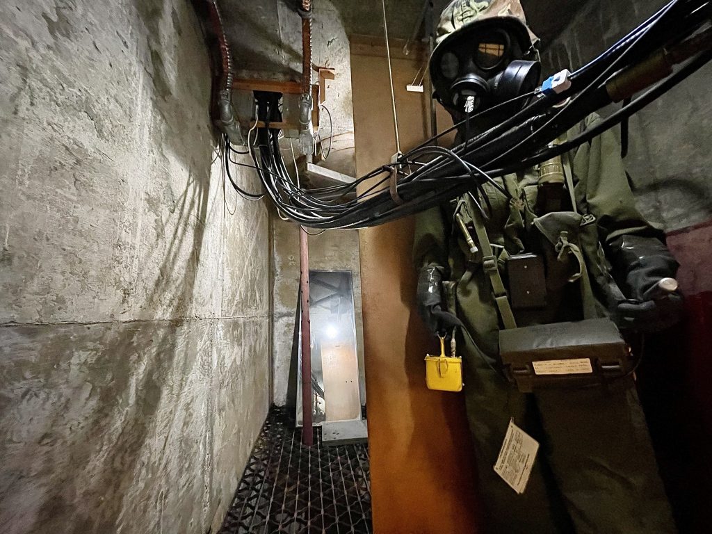 Mannequin dressed in Cold War apparel situated to the right in the Diefenbunker’s escape hatch. A large mass of wires drape in front of the mannequin and continue diagonally across the escape hatch and disappear into the top left corner.