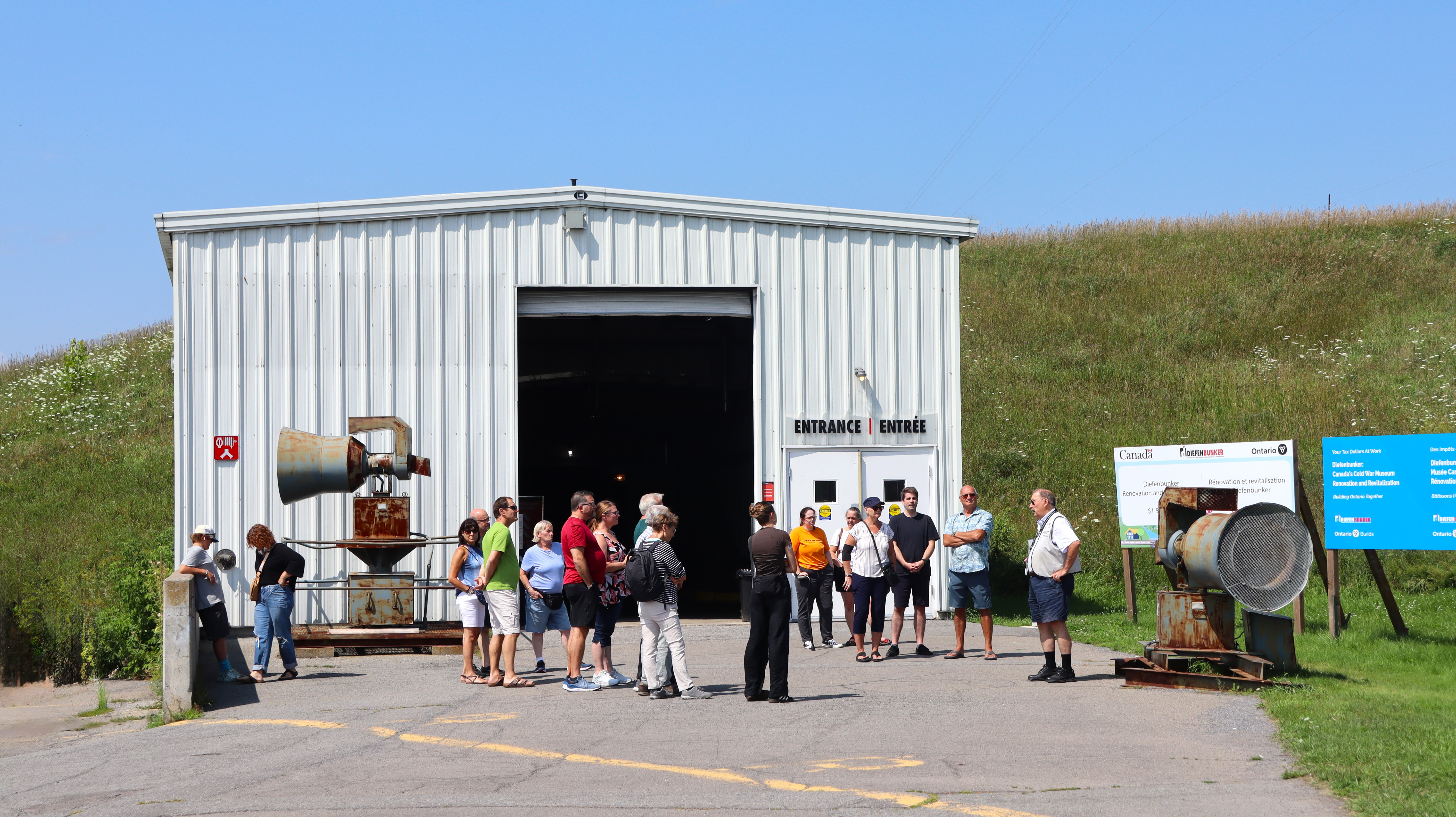 A group of visitors engages with a museum tour guide in front of a white garage-style building that is tucked into a grassy hillside.