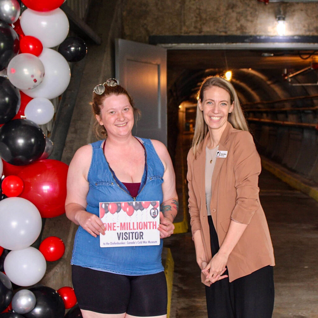 The Diefenbunker's one-millionth visitor holds up a sign, standing beside Diefenbunker Executive Director Christine McGuire, with red, black, and white balloons behind them.
