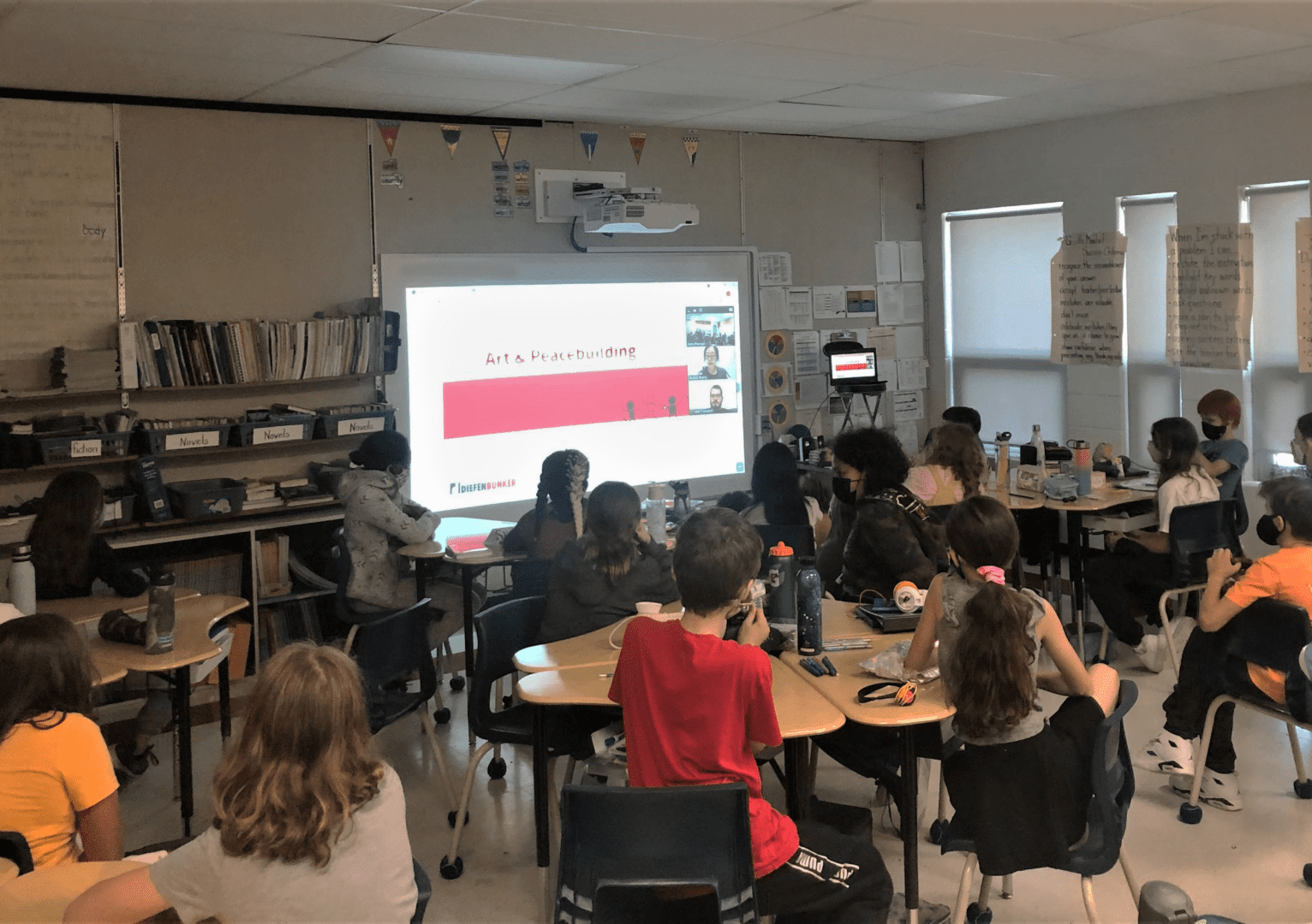 Students in a classroom watch an educational program taking place on the projector screen.