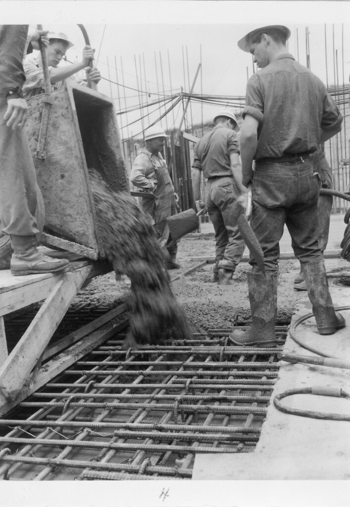 Black and white photograph of workers with a wheelbarrow pouring concrete over rebar.