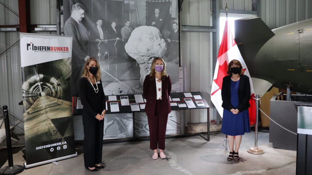 Diefenbunker Executive Director Christine McGuire, MP for Kanata-Carleton Jenna Sudds, and Diefenbunker Board Vice President Joanne Charettestand in front of an exhibit at the entrance to the Diefenbunker.