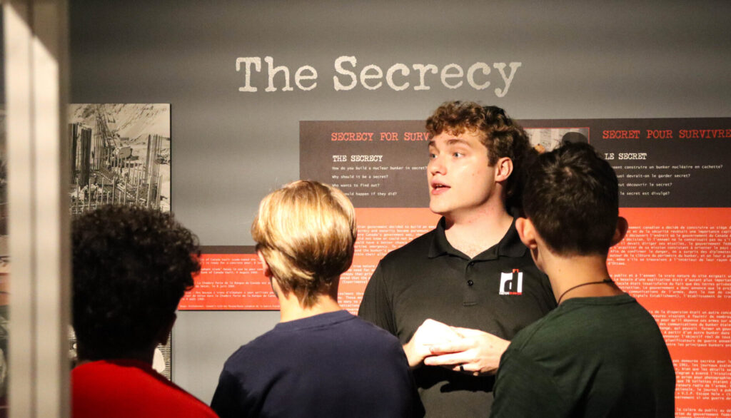 A man stands in front of an exhibition panel that says "The Secrecy" as he speaks to a group of high school students.