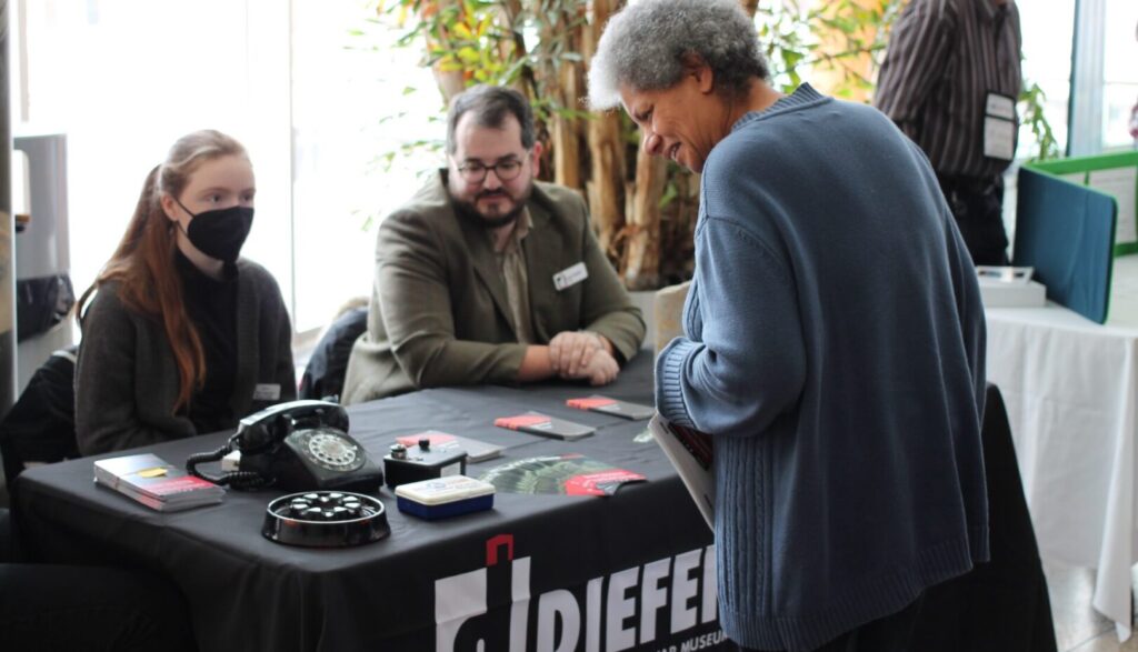 Two people sit behind a table with Diefenbunker brochures and merchandise, and speak to a person standing in front of the table.