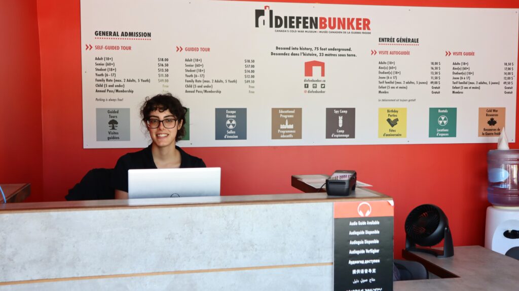 A Diefenbunker staff member smiles in front of the Welcome Desk at the museum.