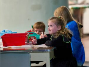 Three young children sit at a table in the Diefenbunker Museum doing a colouring activity.