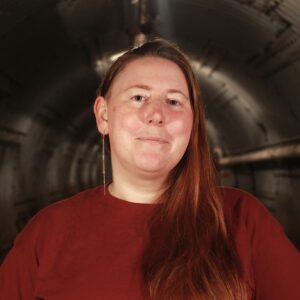 Smiling woman with long red hair in a maroon shirt against a dark background.