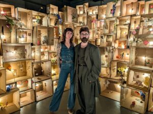 Diefenbunker 2025 Artists-in-Residence Olivia Johnston and Neeko Paluzzi stand in front of one installation in their exhibition, a structure of cardboard boxes, each filled with flowers and light.