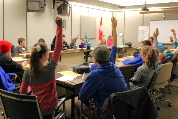 Students sit around a U-shaped table and some of them are raising their hands.