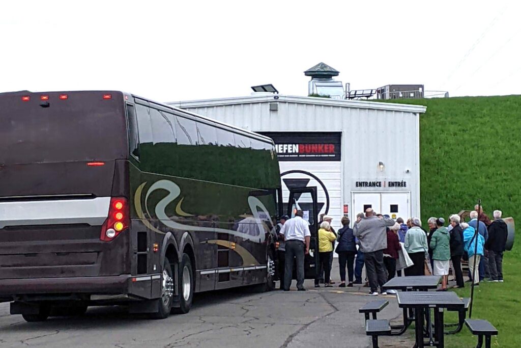 Adults get off a tour bus that is parked in front of a white industrial-style building in front of a grassy hillside.