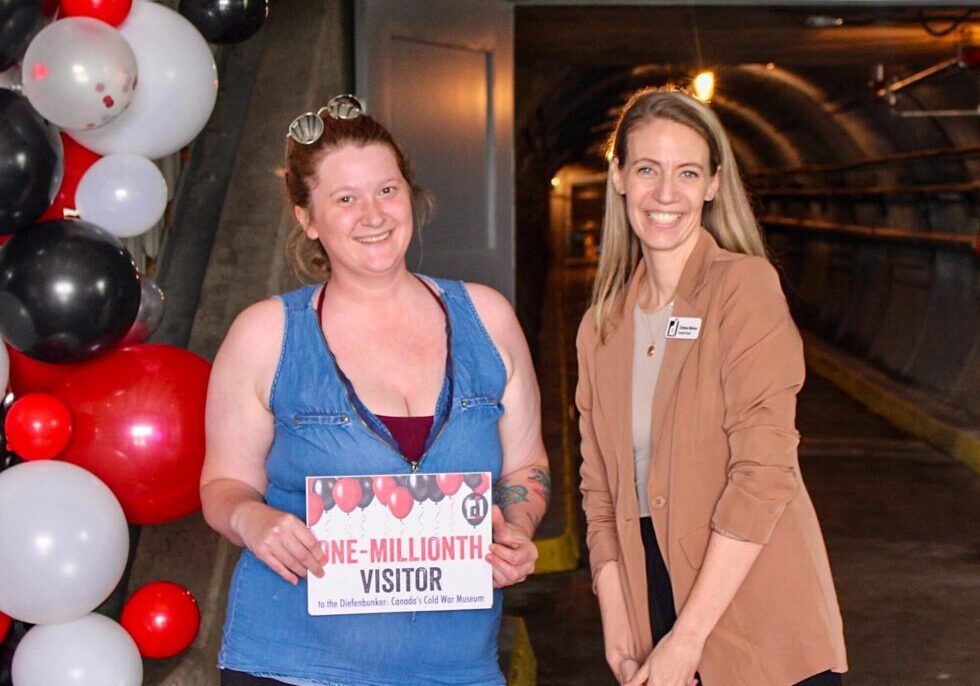 The Diefenbunker's one-millionth visitor holds up a sign, standing beside Diefenbunker Executive Director Christine McGuire, with red, black, and white balloons behind them.