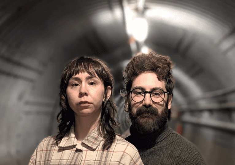 Two people standing in a dimly lit tunnel, one wearing glasses and the other with a serious expression.